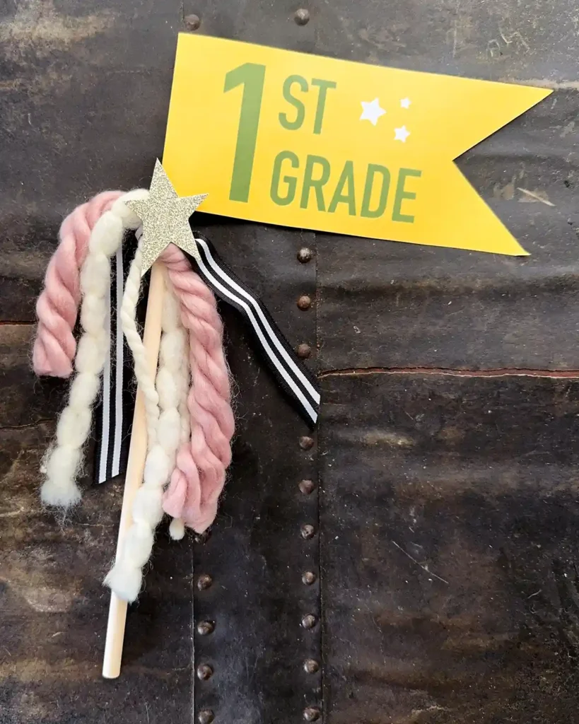 First day of first grade flag made from a yellow paper flag with green font, attached to a wooden dowel, decorated with yarn, ribbon, and a gold star