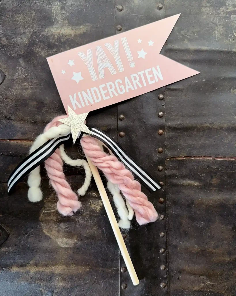 First day of kindergarten flag with white font on a pink background, attached to a wooden dowel decorated with various yarns, ribbons, and a gold glittered star