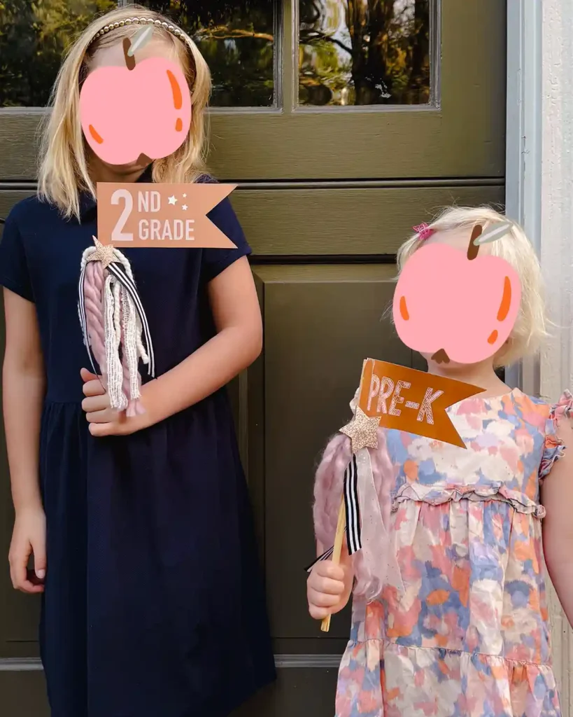 Two girls on their first day of preschool and second grade standing outside the front door holding first day of school flags