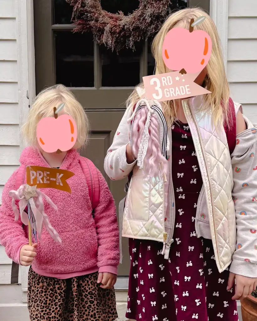 Two girls on their first day of preschool and third grade holding first day of school flags and standing on the deck outside of the house's front door