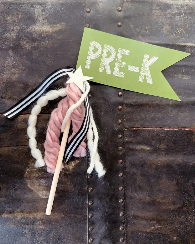 First day of preschool flag with white font on a green background, attached to a wooden dowel decorated with a mix of yarn, ribbon, and a gold glittery star