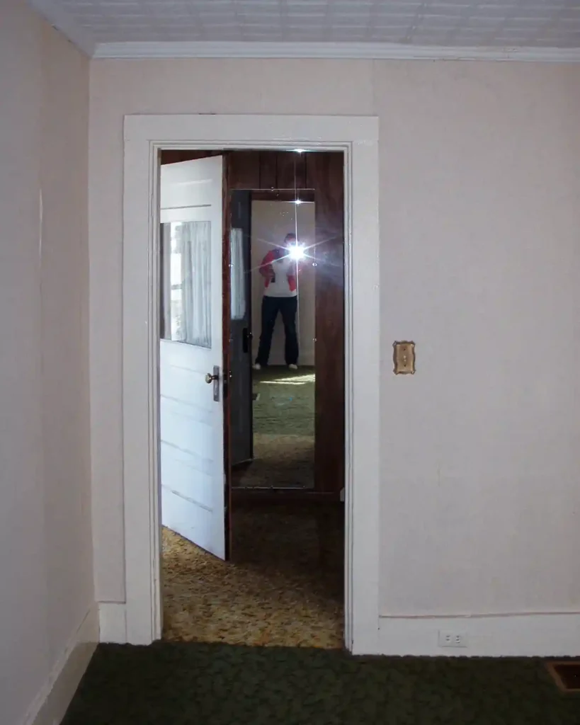 View from a dated living room through a doorway into a hallway at the bottom of the stairs; front door is swung open and a mirror sits behind the door; before photo of the house featuring shag carpeting of various colors and paneled walls