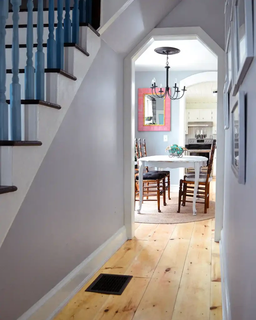 After picture of a renovated hallway featuring newly installed wide pine floors, white trim, greige walls, gallery wall with silver frames, and an updated staircase; view of the hallway from the front door towards the dining room and kitchen