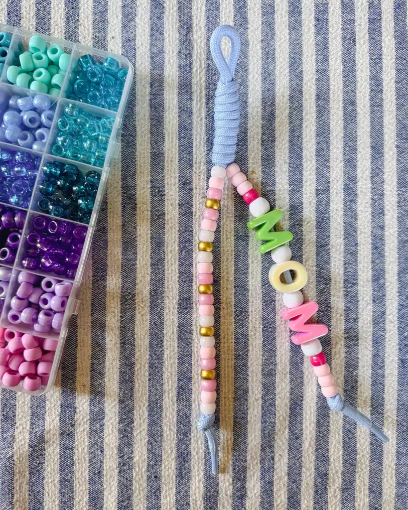 Making paracord bubble letter keychains featuring a light blue cord with beads strung on two lengths with bubble letters spelling mom and a case of pony beads sitting on the side