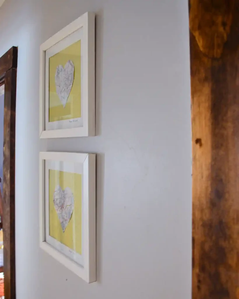 A pair of handmade art with hearts cut out of maps representing places we loved, in white frames hanging in the hallway outside a bedroom door