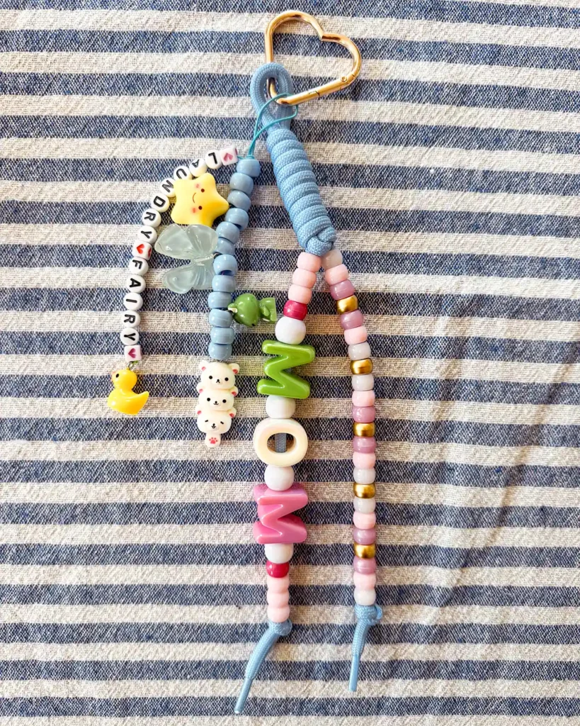 A finished "mom" bubble letter bag charm with blue paracord, a gold heart carabiner, lots of charms and pony beads, and small letter beads that spell "laundry fairy"
