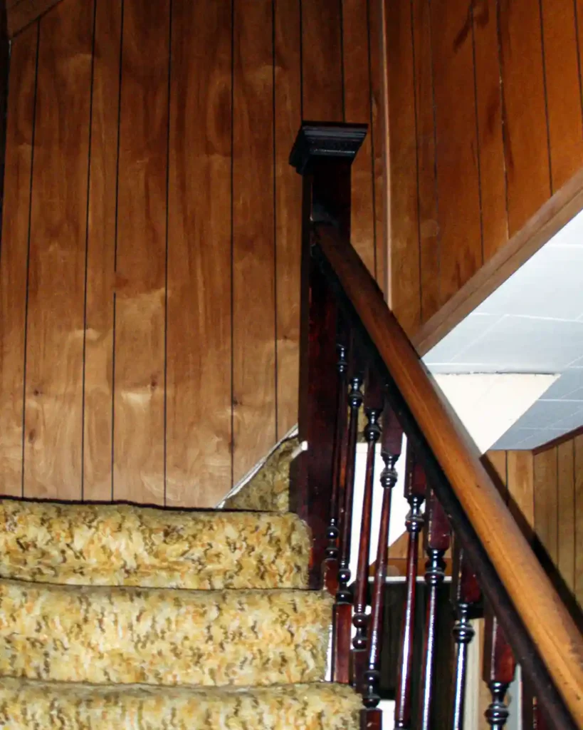 A faux-wood paneled staircase that wraps to the right covered in shag carpeting up the stairs, dark stained balusters and railing, and a dated 1970s style; before picture prior to renovating