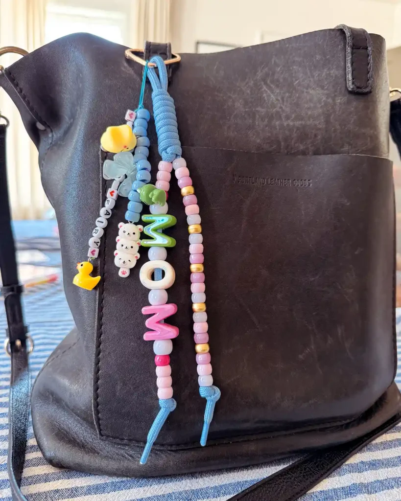 A black leather purse adorned with a chunky letter bead bag charm keychain that says "mom"
