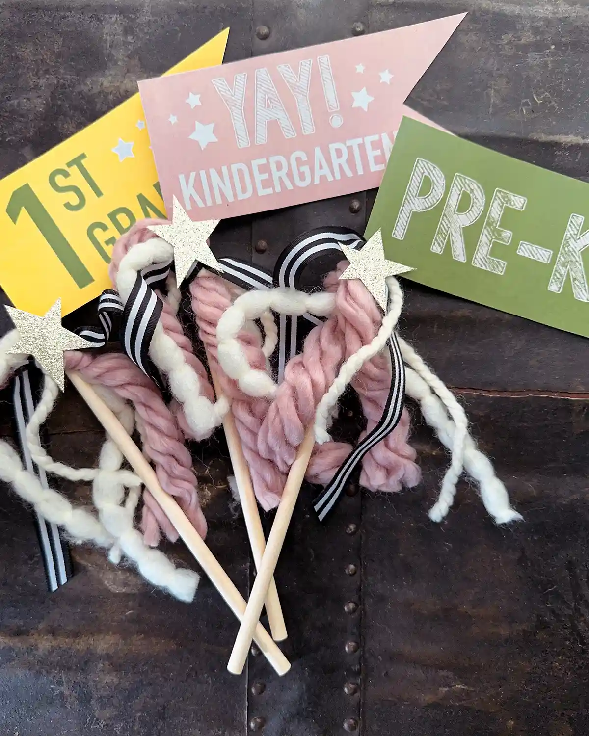 First day of school flags featuring a yellow first grade, pink kindergarten, and green pre-k flags with wooden handles and decorated with yarn and ribbon