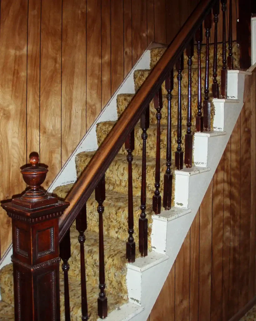 A vintage 1970s style carpeted staircase before tackling a diy staircase renovation; features shag carpeting up the stairs, wood paneled walls, broken balusters, and dark stained newell post