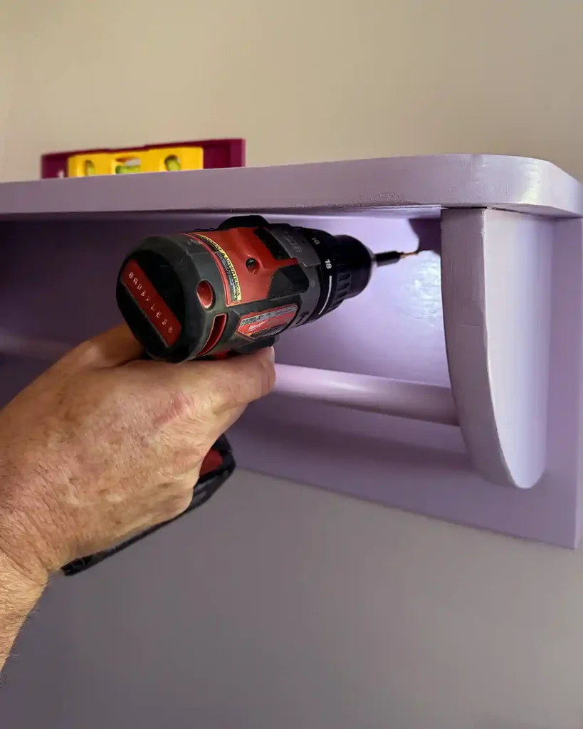 Attaching a purple clothes rack to the wall by drilling through the back of the shelf with a cordless drill