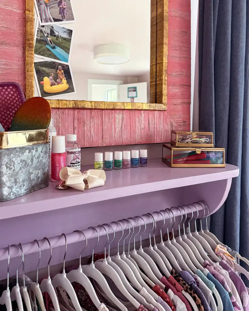 A DIY wooden clothes rack with a shelf on top that's painted purple, kids' dresses hanging on the rack, pink framed mirror above the shelf, hair brushes in a tin, and other kids' items on the shelf like nail polish, body glitter, hair crayons, hair ties and bows, and more