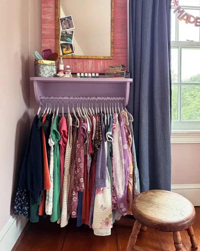 A purple painted DIY wooden clothes rack with a shelf hanging on the wall with girls' dresses hanging below and a pint mirror hanging above the rack