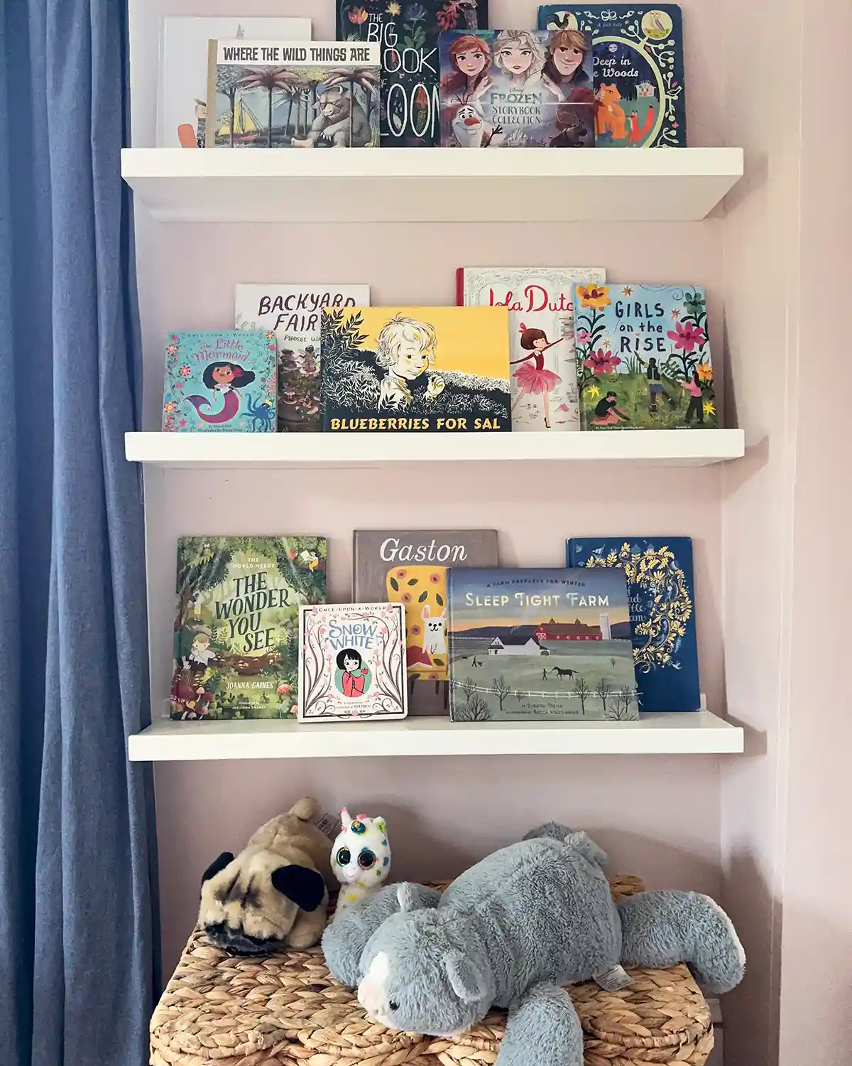 Three DIY ledge shelves painted white and hanging on the wall in a child's bedroom with a variety of picture books and board books arranged on the shelves; a pile of stuffed animals sit on top of a basket below the shelves