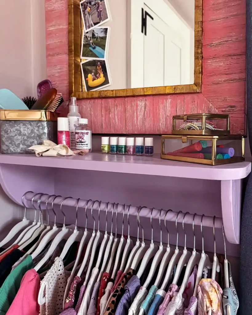 A DIY built and purple painted wall-mounted dress rack for kids' dresses; a collection of clothes hanging from the clothes rack with a shelf above holding all kinds of hair and nail items and a pink framed mirror hanging on the wall above