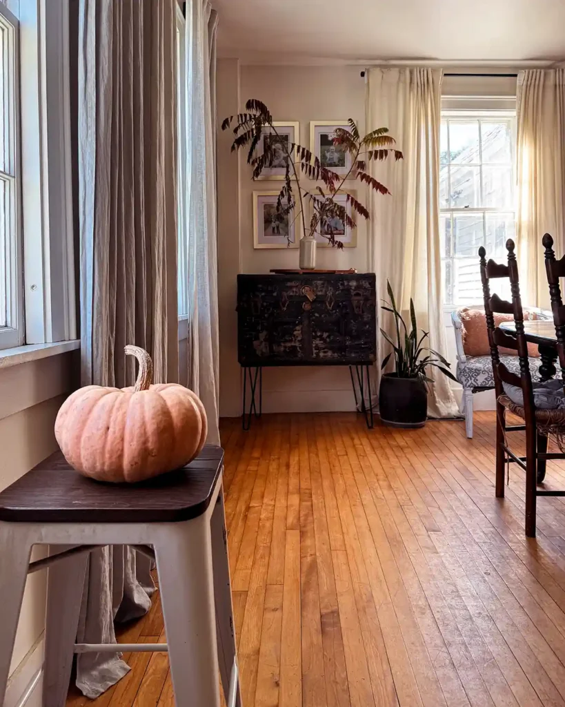 A light and airy dining room decorated for fall with a vase of sumac branches on a side table and a pumpkin sitting on a stool