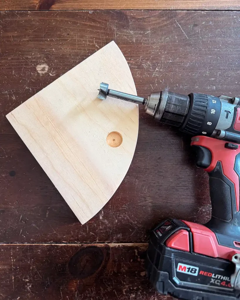 Using a Milwaukee drill with a forstner bit to drill a flat bottomed hole into a quarter round piece of wood that's getting used as a shelf bracket