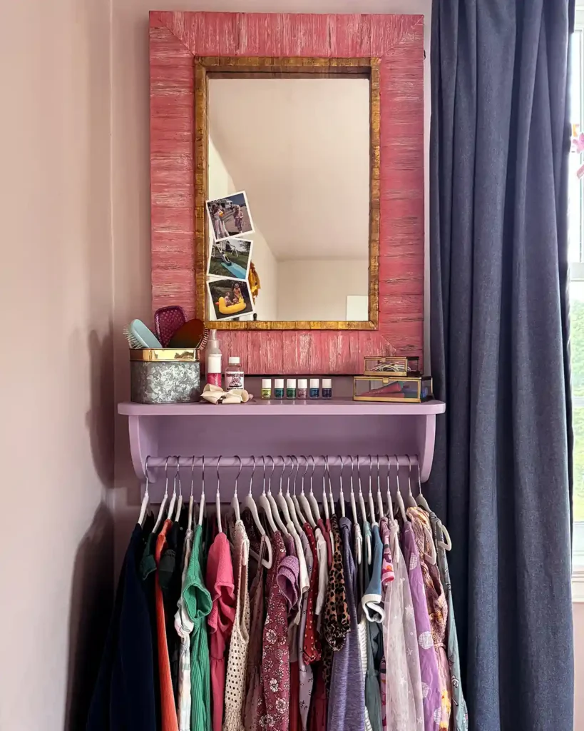 How we built this wall-mounted clothing rack for our daughter's dresses; purple painted rack with a shelf above, pink framed mirror hanging above, collection of dresses hanging from the rack, and hair tools and accessories on the shelf