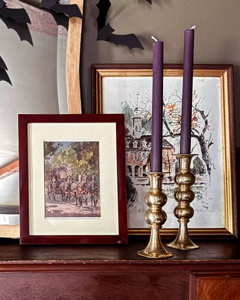 A pair of gold candle holders with ribbed purple tapers sitting on a mantel in front of a pair of art prints and a gold framed mirror