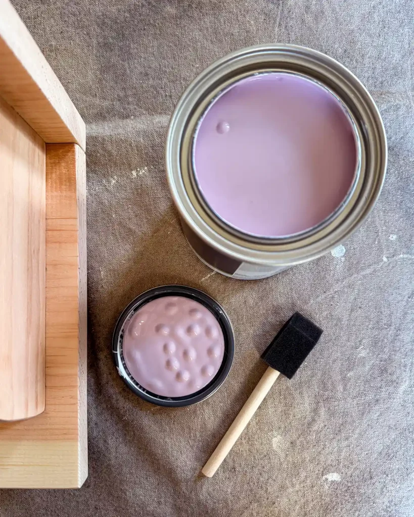 An open paint can of Purple Easter Egg Benjamin Moore paint sitting open on top of a drop cloth next too a wood shelf and foam paint brush