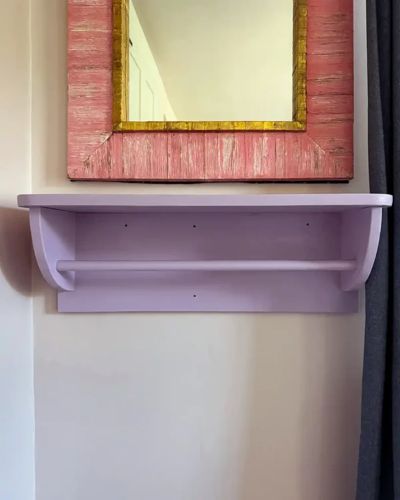 A purple dress rack and shelf attached to the wall with the screw holes visible before filling; pink mirror hanging above the shelf