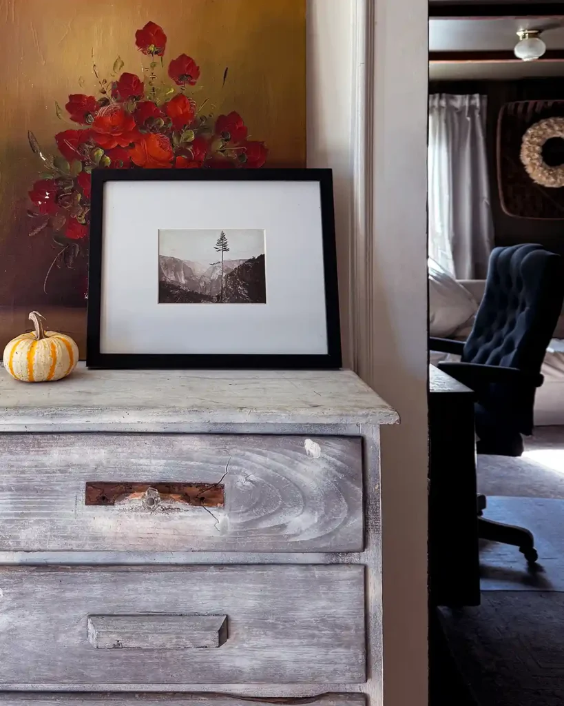 A floral painting and a framed landscape photo sitting on top of a weathered, vintage dresser with a small pumpkin for fall decor