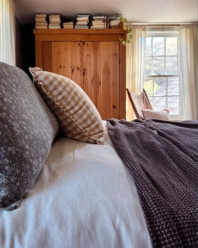 A queen-sized bed updated for fall with soft, blue/green floral flannel sheets and pillow cases, and dark brown coverlet, and a dusty yellow gingham throw pillow; pine wardrobe with books stacked on top in the background