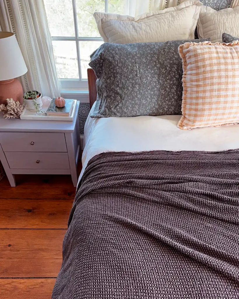 Bedding refreshed for fall with blue floral flannel sheets, white duvet, brown textured coverlet, and a golden yellow gingham throw pillow on the bed; beside the bed is a light colored nightstand with two drawers