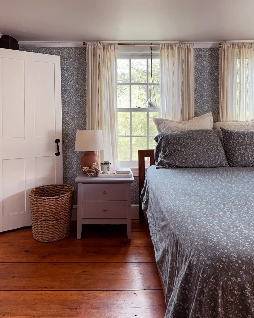 A bed made with a dark, muted blue floral flannel sheet set in front of a pair of windows with a nightstand beside it