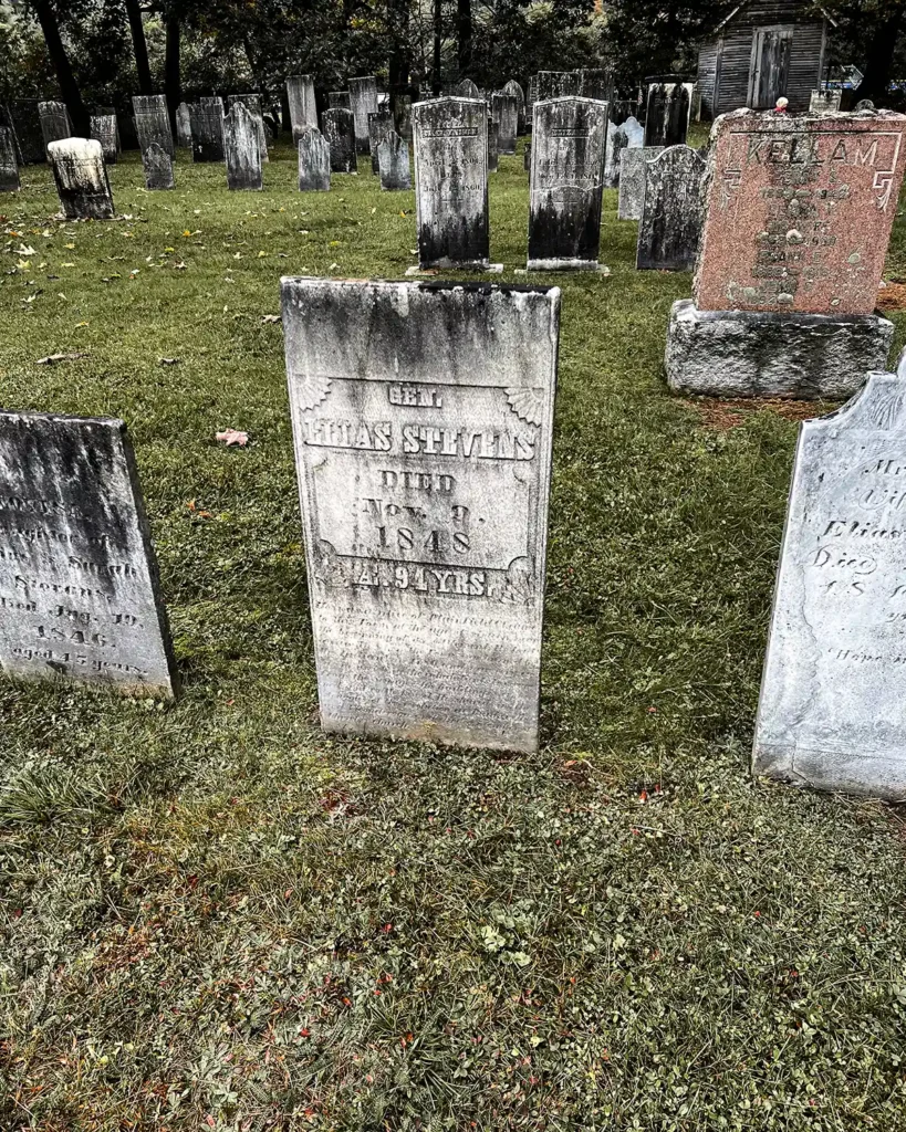 An old cemetery in Vermont filled with weathered gravestones from the 1800s; trees surrounding the cemetery along with a small shed