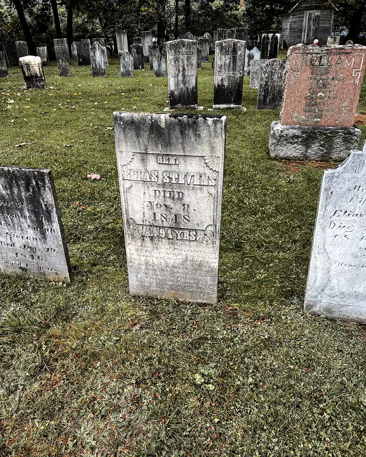 An old cemetery in Vermont filled with weathered gravestones from the 1800s; trees surrounding the cemetery along with a small shed
